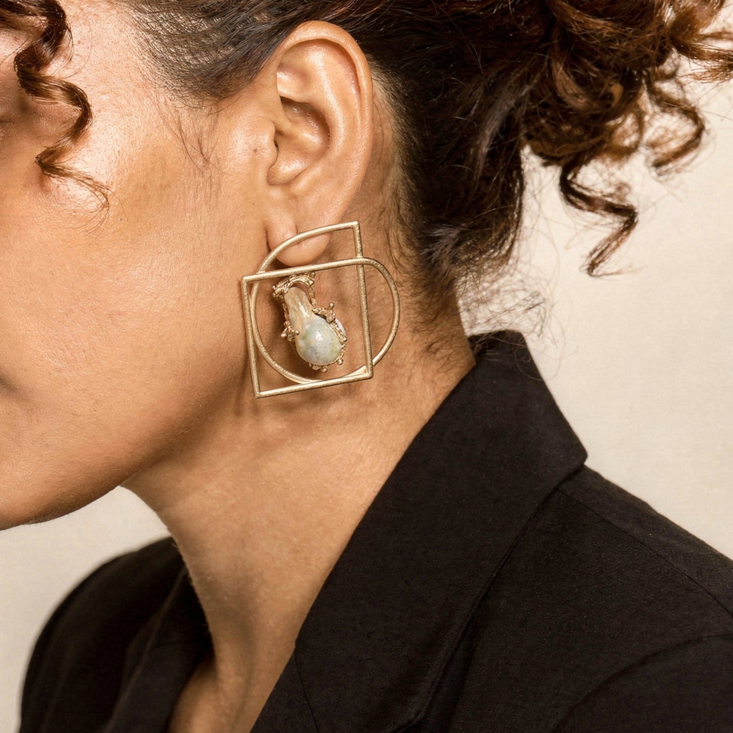 Close-up of a person wearing a gold earring with a central pearl on a neutral background