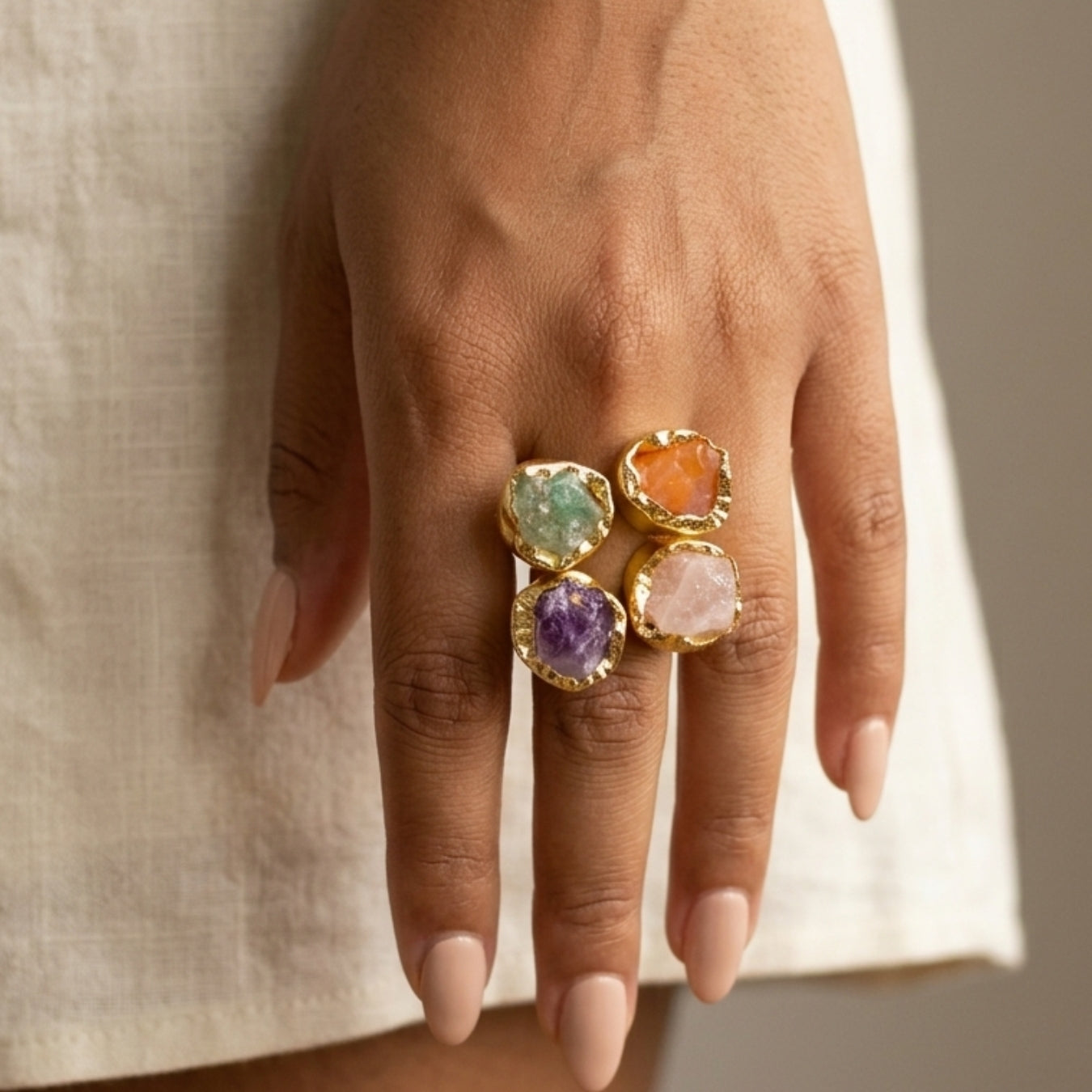 Hand wearing a gold ring with colorful gemstones against a neutral background